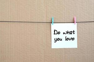 Doen wat u liefde. Notitie is geschreven Aan een wit sticker dat blijft hangen met een wasknijper Aan een touw Aan een achtergrond van bruin karton foto