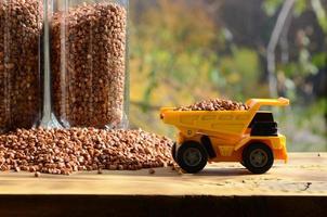 een klein geel speelgoed- vrachtauto is geladen met bruin granen van boekweit in de omgeving van de boekweit stapel en een glas van kruis. een auto Aan een houten oppervlakte tegen een achtergrond van herfst Woud. boekweit levering foto