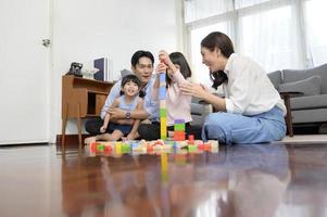 Aziatisch familie met kinderen spelen en gebouw toren van kleurrijk houten speelgoed- blokken in leven kamer Bij huis, leerzaam spel. foto