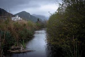 visie van een water kanaal tussen moeras grassen en struiken Bij een regenachtig dag met een berg en een regeling zichtbaar in de afstand foto