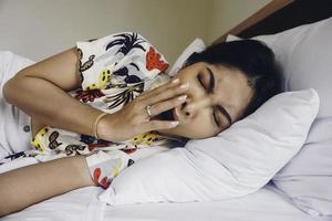 een jong Aziatisch vrouw Aan de bed voelt slaperig, ze is aan het liegen Aan de kant en geeuwen foto