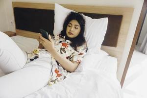 een slaperig jong vrouw geeuwen terwijl Holding telefoon Aan de bed. foto