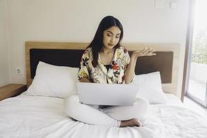 glimlachen jong Aziatisch meisje werken van een afstand achter een laptop Bij huis, zittend in pyjama in bed foto