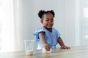 een schattig weinig meisje geniet schoonmaak gemorst melk Aan de tafel. foto
