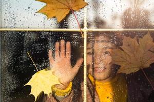 kind looks uit van de venster van de huis buiten, herfst het weer, nat glas met druppels na regenen, geel esdoorn- bladeren zit vast naar de venster. herfst stemming, huis comfort foto