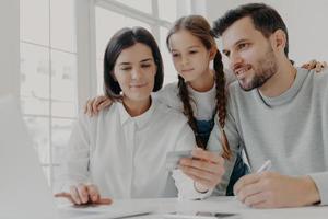 gelukkige vriendelijke familie van vader, moeder en dochter zitten voor de computer, controleer het saldo op de creditcard, doe online winkelen, koop iets dat nodig is, gebruik een snelle internetverbinding. foto