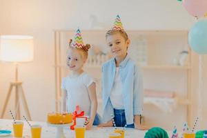 indoor shot van klein meisje en jongen dragen feestmutsen, staan in de buurt van feestelijke tafel met cake, papieren bekers en cadeau, vieren samen verjaardag, poseren in witte ruime kamer. viering concept. foto