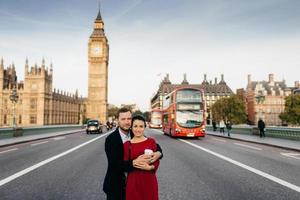 buitenshuis schot van romantisch paar omhelzing elk ander, staan Aan Westminster brug, hebben excursie, houding Bij camera tegen groot ben en bezig stad tafereel met vervoer, genieten wezen in Londen foto