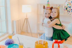 vriendelijke twee meisjes omhelzen en hebben een goede relatie, staan in de buurt van een feestelijke tafel met cake, vieren samen verjaardag, staan in de woonkamer. blije vrouwelijke zussen genieten van vakantie, speciale gelegenheid foto