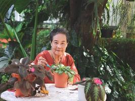 gelukkig en gezond Aziatisch senior vrouw zittend Bij wit tafel buitenshuis nemen zorg van planten, glimlachen en op zoek Bij camera. foto