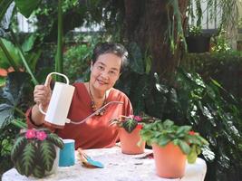 gelukkig en gezond Aziatisch senior vrouw zittend Bij wit tafel buitenshuis nemen zorg van planten. gieter episch cupreata of vlam paars in fabriek pot. op zoek Bij camera. foto