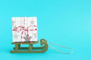 geschenk doos verpakt van wit en grijs papier met rood Wit lint Aan een groen houten slee Aan een helder blauw achtergrond. Kerstmis en nieuw jaar concept met kopiëren ruimte. foto