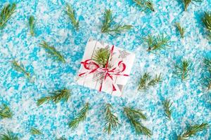 klein geschenk doos verpakt van wit en grijs papier met rood Wit lint Aan de blauw achtergrond met kunstmatig sneeuw versierd Spar takken, top visie. Kerstmis en nieuw jaar concept met kopiëren ruimte. foto