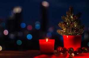 brandend kaars in rood glas met Kerstmis boom en snuisterij ornamenten Aan kleurrijk stad bokeh licht achtergrond. foto