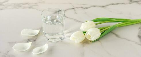 een glas van gezuiverd vers drinken water Aan een marmeren tafel met wit tulp bloemen . kopiëren ruimte voor tekst foto