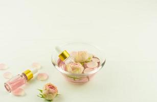 twee glas kunstmatig flessen met roos olie, bloemknoppen en roos bloemblaadjes in een glas kom Aan een wit tafel. foto