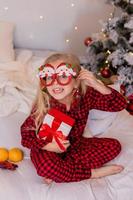 gelukkig weinig blond meisje Bij huis in pyjama voor Kerstmis sorteren presenteert en spelen met Kerstmis speelgoed foto