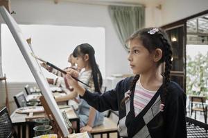 een weinig meisje concentraten Aan acryl kleur afbeelding schilderij Aan canvas met multiraciaal kinderen in een kunst klas, creatief aan het leren met talenten en vaardigheden in de elementair school- studio onderwijs. foto