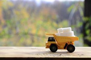 een klein geel speelgoed- vrachtauto is geladen met wit suiker kubussen. een auto Aan een houten oppervlakte tegen een achtergrond van herfst Woud. extractie en vervoer van suiker foto