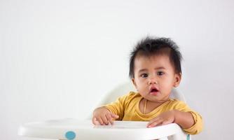 baby meisje van Aziatisch etniciteit geel overhemd zit geduldig in een stoel alleen Bij huis. uitdrukking van emotie, spanning, jeugd, en kinderachtigheid. uit dat schattig Wauw gezicht geluk leven foto