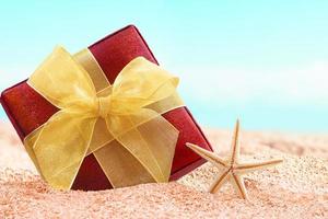 rood geschenk doos met geel boog Aan zand strand met zeester. uitverkoop. kerstmis, nieuw jaar. vakantie in heet landen foto