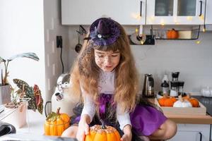 kind siert de keuken in huis voor halloween. meisje in een heks kostuum Toneelstukken met de decor voor de vakantie - vleermuizen, jack lantaarn, pompoenen. herfst comfort in huis, scandi-stijl keuken, zolder foto