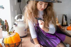 kind siert de keuken in huis voor halloween. meisje in een heks kostuum Toneelstukken met de decor voor de vakantie - vleermuizen, jack lantaarn, pompoenen. herfst comfort in huis, scandi-stijl keuken, zolder foto