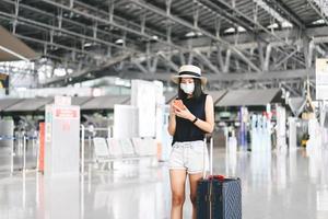 volwassen toerist vrouw met bagage slijtage masker voor covid-19 controleren in met mobiel telefoon Bij luchthaven terminal. foto