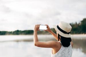 achterzijde visie van jong volwassen Aziatisch vrouw gebruik makend van mobiel telefoon mockup blanco wit scherm Bij strand Aan dag. foto