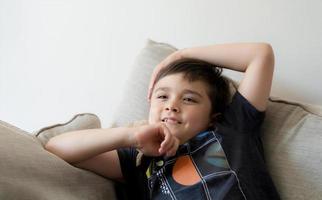 portret gelukkig kind lachend, schattig weinig jongen met groot glimlach zittend Aan bankstel hebben pret aan het kijken tekenfilm Aan TV, positief kind met glimlachen gezicht ontspannende genieten gelukkig tijd alleen Aan weekend. foto