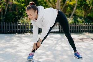 gemotiveerde jongvolwassen vrouw gaat regelmatig sporten reikt regelmatig hand aan voet leunt naar beneden heeft voeten op schouderbreedte uit elkaar draagt sportkleding vindt het leuk om buitensporten te trainen om af te vallen foto