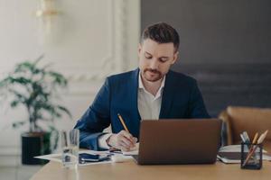 serieuze geconcentreerde geschoolde mannelijke ondernemer controleert financiële boekhouding rapport schrijft enkele notities in notitieblok zit in coworking space gebruikt laptopcomputer formeel gekleed. bezettingsconcept foto