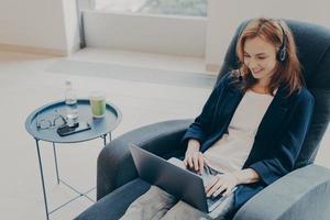 jonge ondernemersdame met een hoofdtelefoon die een laptop gebruikt terwijl ze thuis in een fauteuil zit foto