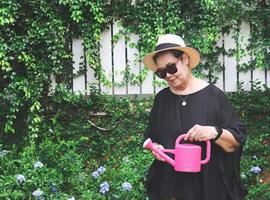 gelukkig en gezond senior Aziatisch vrouw vervelend zwart blouse, zonnebril en hoed, Holding roze gieter kan in de tuin, glimlachen en op zoek Bij gieter kan. foto