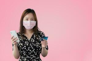 gelukkig Aziatisch portret schattig jong vrouw vervelend chirurgisch masker houden mobiel telefoon en plastic debiteren credit bank kaart, studio schot geïsoleerd Aan roze achtergrond, vrouw gebruik makend van smartphone online boodschappen doen foto