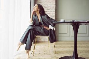 ballerina in een grijs gestreept pak met een kop van koffie glimlachen en dromen is zittend Bij een tafel door de venster foto