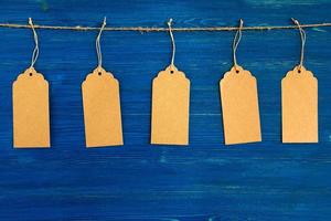 vijf bruin blanco papier prijs tags of etiketten reeks hangende Aan een touw Aan de blauw houten achtergrond. foto