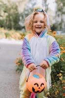 schattig weinig meisje in een regenboog eenhoorn kostuum voor halloween gaat naar verzamelen snoepgoed in een pompoen mand in een woon- Oppervlakte. kind wandelingen in de buitenshuis. truc of traktatie. levensstijl. kigurumi foto
