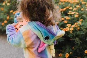 schattig weinig meisje in een regenboog eenhoorn kostuum voor halloween gaat naar verzamelen snoepgoed in een pompoen mand in een woon- Oppervlakte. kind wandelingen in de buitenshuis. truc of traktatie. levensstijl. kigurumi foto