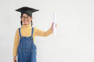 Aziatisch weinig meisje vervelend een diploma uitreiking pet en Holding diploma Aan wit achtergrond foto