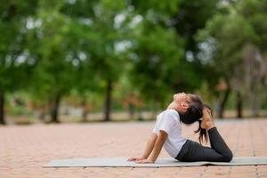 weinig schattig Aziatisch meisje beoefenen yoga houding Aan een mat in park, gezond en oefening concept foto