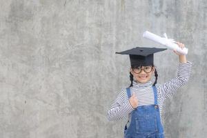 Aziatisch weinig meisje vervelend een diploma uitreiking pet en Holding diploma Aan wit achtergrond foto