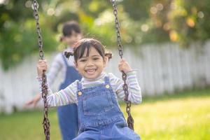 twee schattig weinig zussen hebben pret Aan een schommel samen in een mooi zomer tuin Aan een warm en zonnig dag buitenshuis. actief zomer vrije tijd voor kinderen. foto