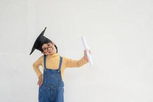 Aziatisch weinig meisje vervelend een diploma uitreiking pet en Holding diploma Aan wit achtergrond foto