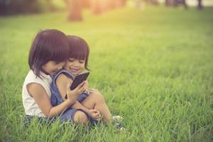 twee kleine zusmeisjes die internet spelen met mobiele smartphone op gras foto