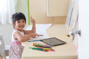 weinig Aziatisch kind gebruik makend van een potlood naar schrijven Aan notitieboekje Bij de bureau foto