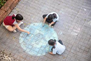 Aziatisch kinderen Speel buitenshuis. kind meisje trekt een planeet wereldbol met een kaart van de wereld gekleurde krijt Aan de stoep, asfalt. aarde, vrede dag concert. foto
