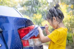 gelukkig Aziatisch meisje het wassen auto Aan water spatten en zonlicht Bij huis foto