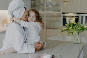 aanhankelijke moeder draagt badjas gewikkelde badhanddoek op hoofd omhelst en kust met liefde haar kleine dochter poseert samen thuis tegen een gezellig interieur voel verfrist na het douchen foto