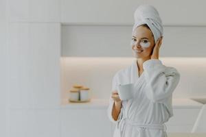 mooie jonge vrouw met een gezonde huid brengt cosmetische pleisters aan onder de ogen drinkt hete thee of koffie uit een mok gekleed in een badjas na het nemen van een douche poseert binnen tegen wit keukenmeubilair foto